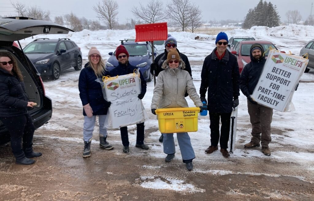 A group of picketers at a picket line holding picket signs, a tote, and a shovel. 